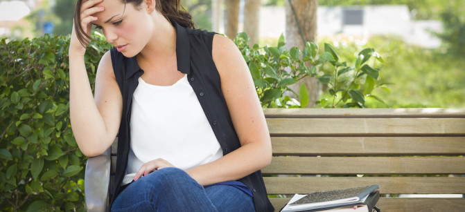 Upset Young Woman Sitting Alone with Her Head in Her Hands on Bench Next to Books and Backpack.