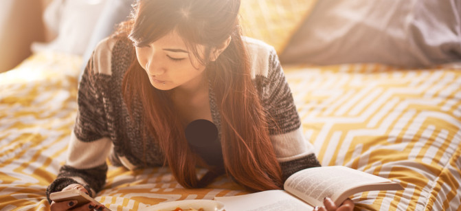 asian teen on bed checking smartphone while reading book and eating soup