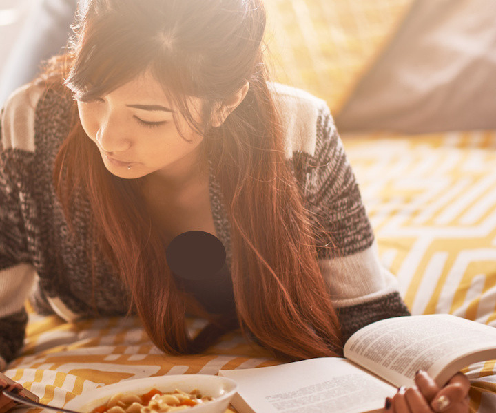 asian teen on bed checking smartphone while reading book and eating soup