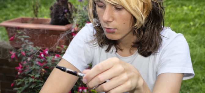 A teenage boy sitting in a garden, smoking an electronic cigarette and exhaling vapour