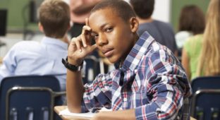Bored Male Teenage Pupil In Classroom Looking At Camera Leaning On Arm