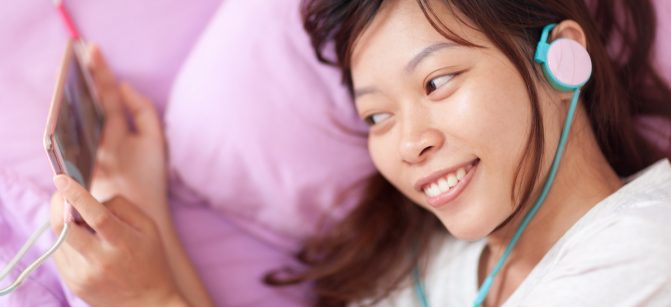 Asian young girl watching mobile video smart phone wear headphones lying on bed top angle view