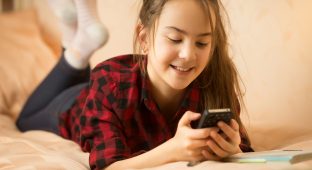 Portrait of smiling teenage girl ling on bed and using smartphone