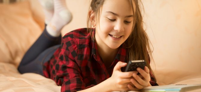 Portrait of smiling teenage girl ling on bed and using smartphone