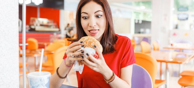 Pretty young funny woman eating hamburger inside in fast-food restaurant