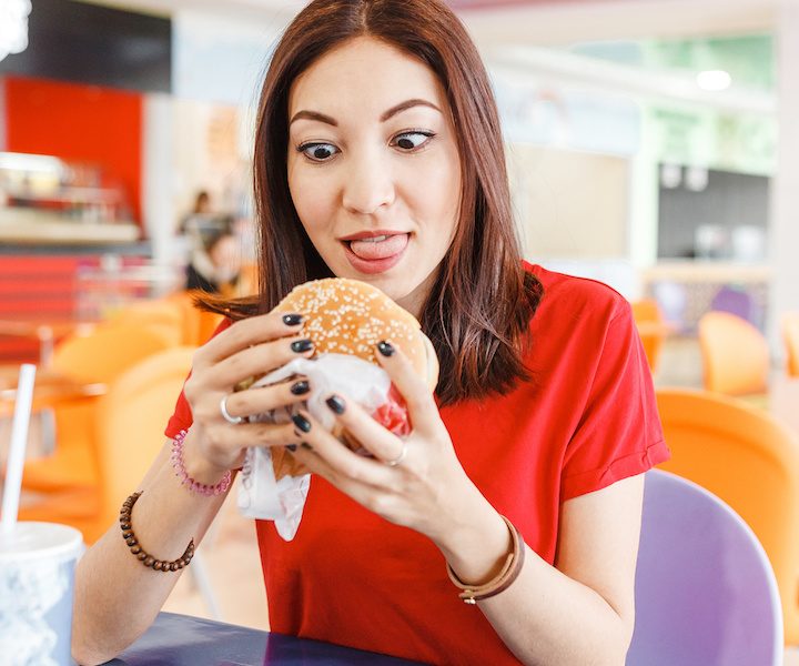 Pretty young funny woman eating hamburger inside in fast-food restaurant