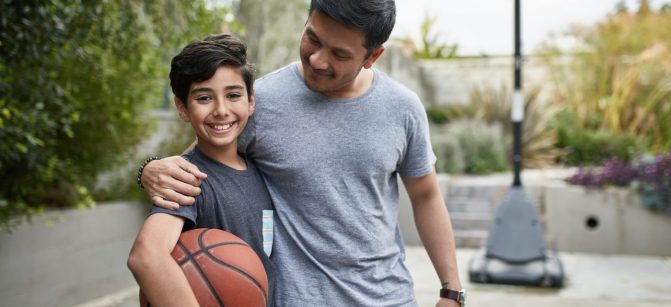 Portrait of happy boy standing with father at basketball court. Mid adult man and child are smiling in backyard. They are in casuals during weekend.
