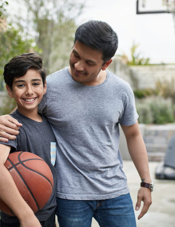 Portrait of happy boy standing with father at basketball court. Mid adult man and child are smiling in backyard. They are in casuals during weekend.