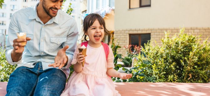 Image of happy cute little girl sitting with dad on the city street and eating ice-cream outdoor. Fun girl kid and father have fun and playing outside. Good relationship between dad and daughter