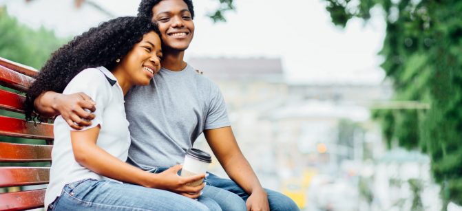 Young african american couple in love hugging, kissing and sitting on the red bench in summer time. Love, holiday, travel and relationship concept