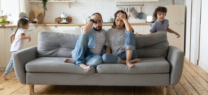 Tired mother and father sitting on couch feels annoyed exhausted while noisy little daughter and son shouting run around sofa where parents resting. Too active hyperactive kids, need repose concept