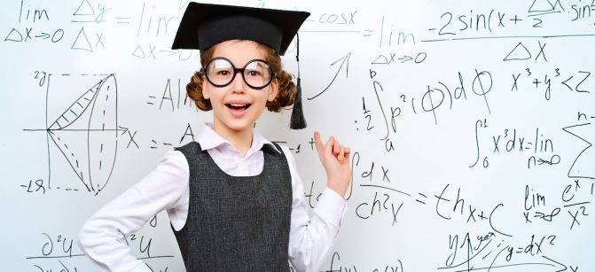 Happy smart schoolgirl in big glasses and academic hat performs the task at the blackboard. Education.