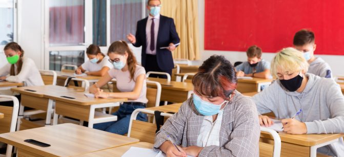 Focused teenage students in protective face masks studying in classroom with teacher, writing lectures in workbooks. Necessary precautions in coronavirus pandemic