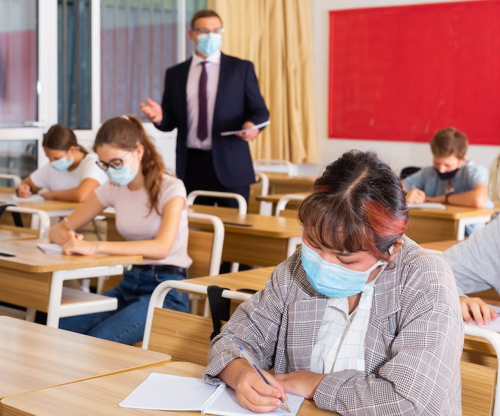 Focused teenage students in protective face masks studying in classroom with teacher, writing lectures in workbooks. Necessary precautions in coronavirus pandemic