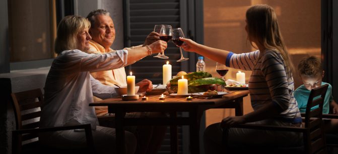 Family dinner outdoor in the backyard in quiet evening. Young woman and senior parents toasting with wine while child playing games