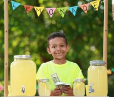 Cute little African-American boy with money at lemonade stand in park. Summer refreshing natural drink