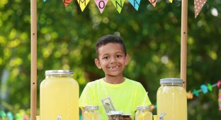 Cute little African-American boy with money at lemonade stand in park. Summer refreshing natural drink