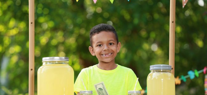 Cute little African-American boy with money at lemonade stand in park. Summer refreshing natural drink