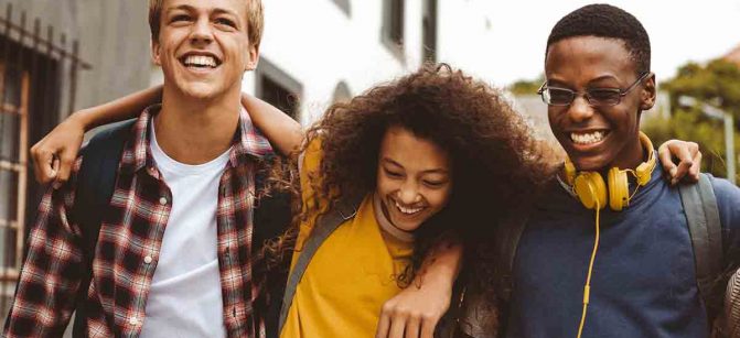 Close up of three college friends standing in the street with arms around each other. Cheerful boys and a girl wearing college bags having fun walking outdoors.