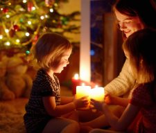 Young mother and her two little daughters sitting by a fireplace holding candles in a cozy dark living room on Christmas eve